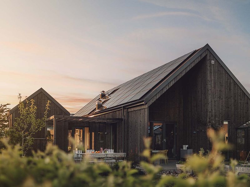 Ein Holzhaus mit Solaranlagen auf dem Dach. Im Hintergrund ist ein Sonnenuntergang zu sehen.