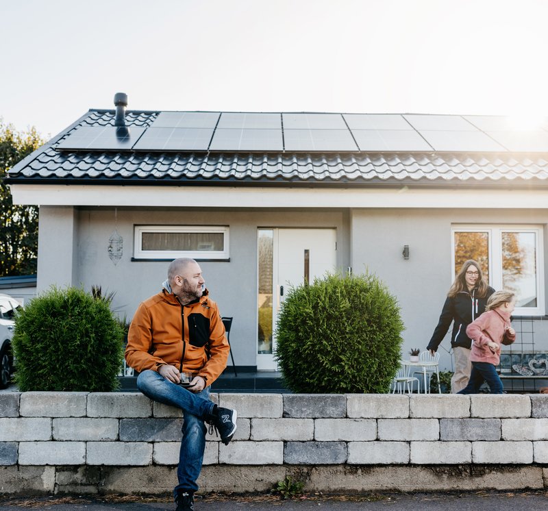 Familie vor ihrem Haus mit einer Photovoltaikanlage. Die Kinder spielen im Vorgarten. (Vorteile und Nachteile von einer Photovoltaik-Anlage).